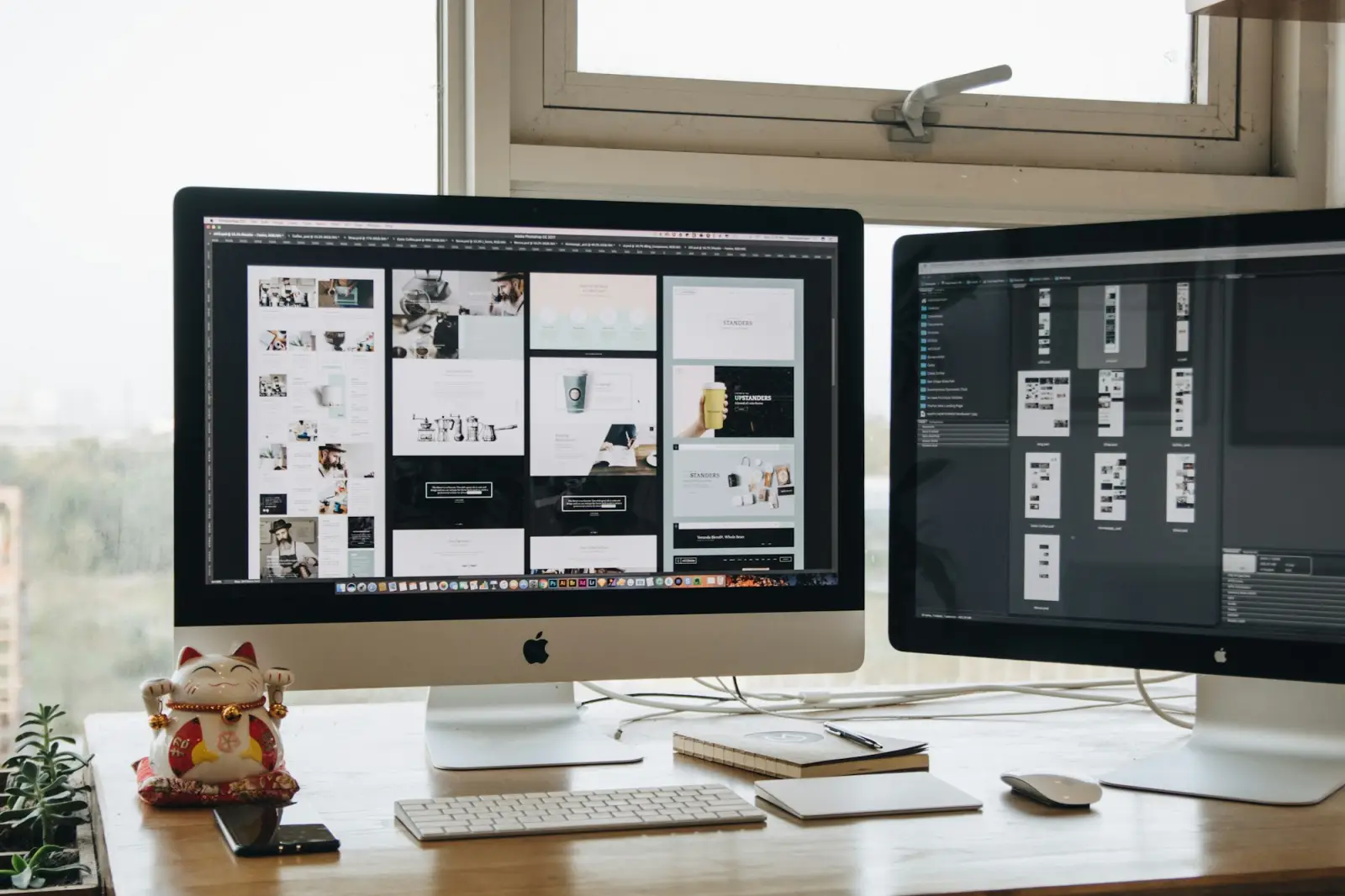 Two desktop monitors showing design layouts on a desk by a window