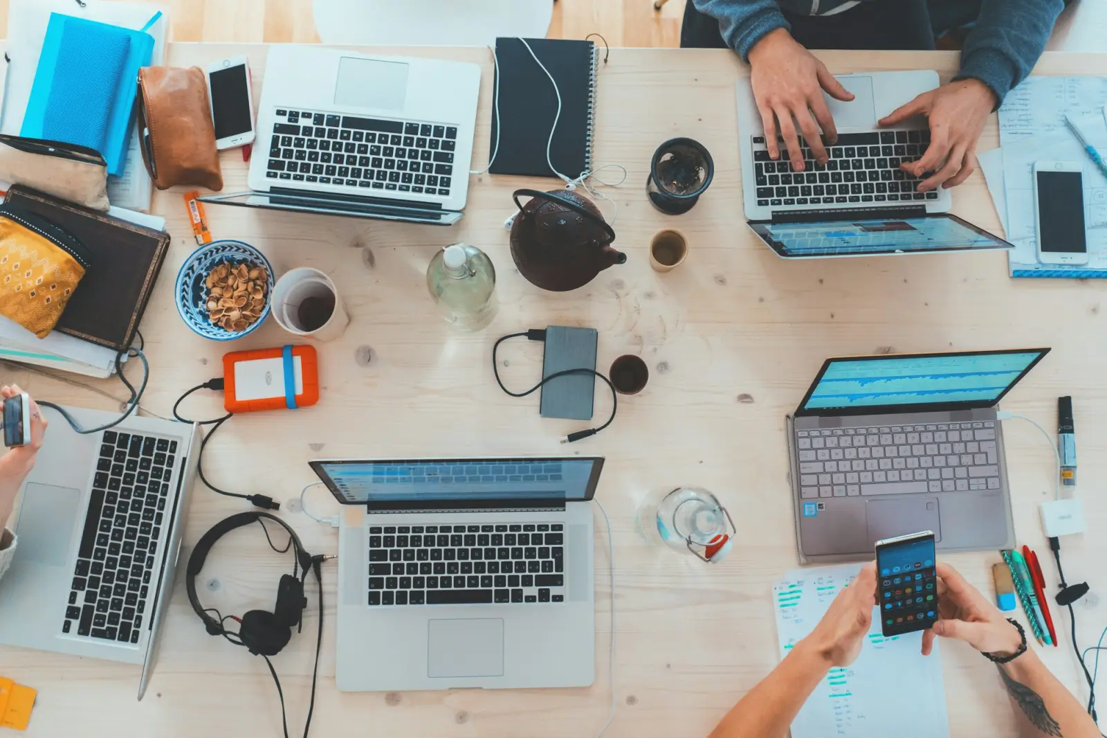Overhead view of a shared table with several laptops, notebooks and hands at work