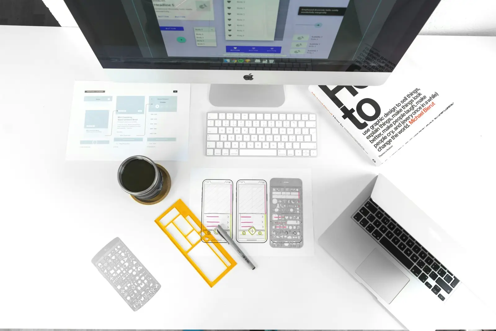 Overhead view of a desk with a desktop computer, a laptop, wireframe sketches and stencils